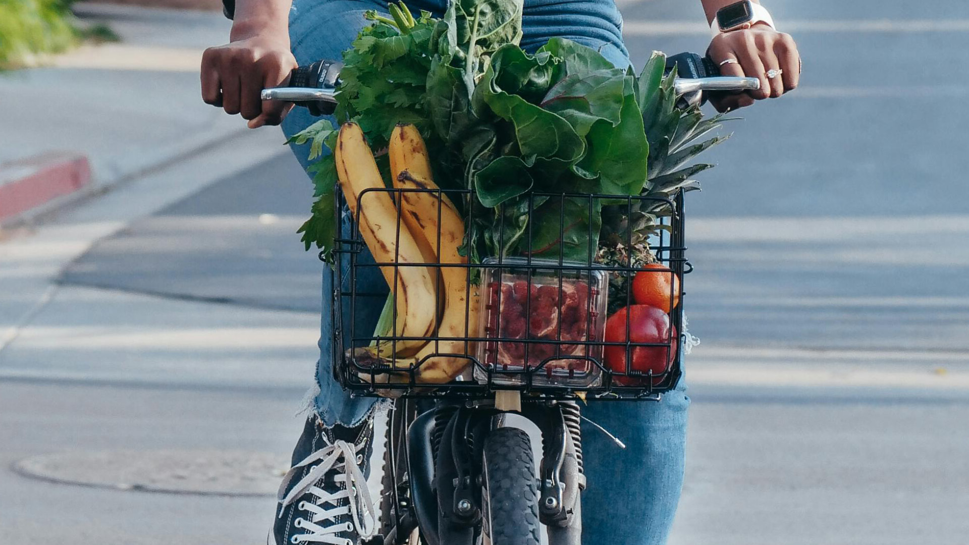 A basket attached to the front of a bicycle, filled with plantain, apples, oranges, pineapple and spinach.